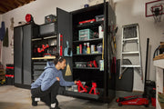 A person is organizing tools and supplies in a Craftsman storage cabinet in a garage. The cabinet contains various items such as cleaning products, bins, power tools, and jack stands. Nearby, there is a ladder, a floor jack, and other garage equipment.